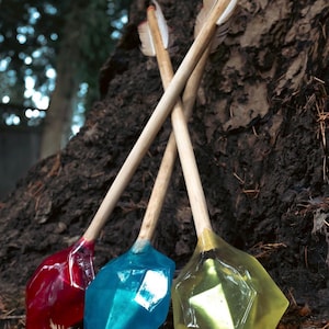 May include: Three arrows with wooden shafts and fletching, each tipped with a large, faceted, translucent resin arrowhead. The arrowheads are coloured in deep red, bright blue, and pale yellow, reflecting light on their angular surfaces.