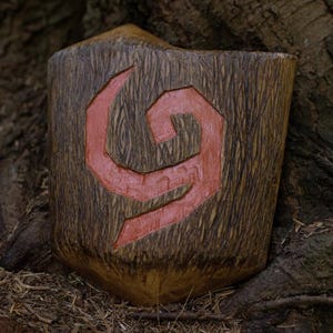 May include: A wooden shield featuring a carved red symbol. The shield has a natural wood grain texture and a rounded shape. The background shows tree bark and foliage, suggesting an outdoor setting.
