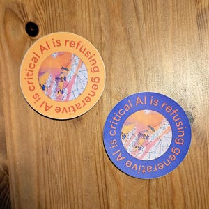 May include: Two round stickers with the text "AI is critical, refusing generative AI". One sticker is orange with a colorful abstract design, the other is blue. Both are on a wooden surface.
