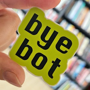 May include: A lime green sticker with the words "bye bot" in bold black letters. The sticker has a rounded, irregular shape. The background is blurred, suggesting it is being held up in front of a shelf.