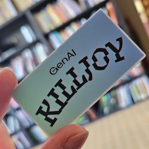 May include: A rectangular card with the text "GenAI KILLJOY" in black, stylized letters. The card has a gradient background of blue and green. The card is held in a person's hand, with a blurred bookshelf in the background.