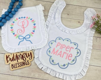 May include: White baby bibs with ruffled edges. One bib features a floral wreath with a pink letter and a blue bow. The other bib has the name "Piper Marie" embroidered in pink and blue. A wooden sign reads "Butterfly Blessings".