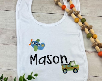 May include: White baby bib with embroidered airplane, truck, and the name "Mason" in black. The bib has a rounded shape and a curved neck opening. A wooden bead necklace is next to the bib.