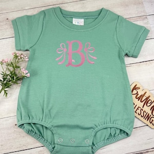 May include: A sage green short-sleeve romper featuring a pink embroidered letter B flanked by two decorative bows. The garment has a round neckline, a tag labeled LTC at the collar, and three metal snap closures at the bottom leg opening.