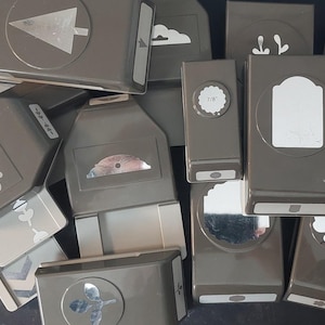 May include: A collection of grey plastic craft punches featuring various shapes including a tree, cloud, cupcake, flower, and leaf. Each tool has a lever mechanism for cutting paper designs, with some units displaying specific size labels like 7/8 inch.