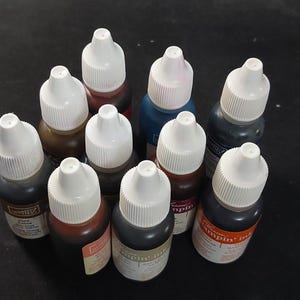 May include: A collection of ten small plastic bottles of Stampin' Ink, each featuring a white pointed applicator cap. The bottles contain various colored inks, including brown, blue, black, and orange, with labels displaying the brand name and color.