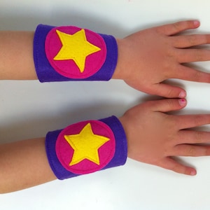 May include: Two purple felt arm bands with pink circles and yellow star appliques. The arm bands are worn on a child's arms.