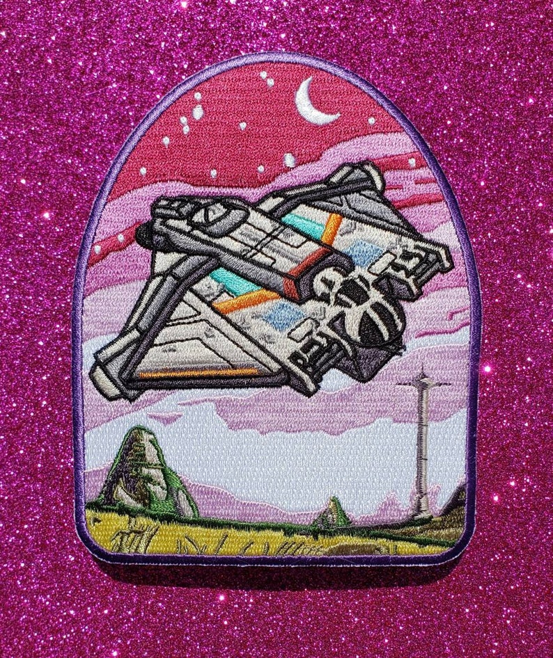 May include: Embroidered patch featuring a white spaceship flying through a pink and purple sky with a crescent moon and stars. The spaceship has orange and blue accents. The background includes green hills and a tall, thin tower.