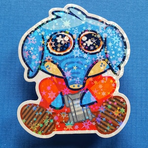 May include: A holographic sticker of a cartoon elephant with a blue head and body, orange arms, and brown legs. The elephant has large eyes and is decorated with star-shaped sparkles. The sticker is on a blue background.