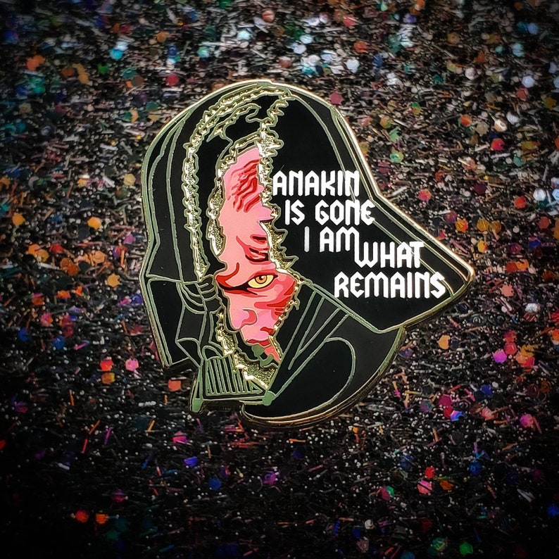 May include: A black and gold enamel pin featuring a split design of Darth Vader's helmet revealing Anakin's face. The pin includes the text "ANAKIN IS GONE I AM WHAT REMAINS" in white lettering. The background is a glittery, dark surface.