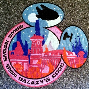 May include: A pink and blue Mickey Mouse-shaped patch with a Star Wars theme. The patch features a cityscape with buildings from Star Wars: Galaxy's Edge, including the Millennium Falcon and TIE fighters. The text on the patch reads "Galaxy's Edge from a galaxy far, far away."