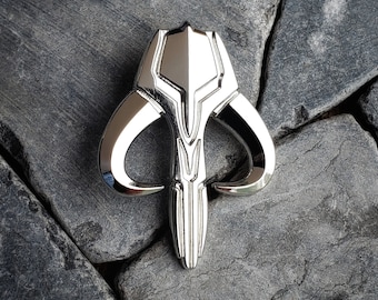 May include: A shiny silver Mandalorian skull emblem pin, featuring a stylized design with curved horns and a central crest. The pin is set against a backdrop of gray stones, creating a striking contrast.