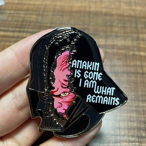 May include: A black enamel pin featuring Darth Vader's helmet with a split revealing Anakin's face. The text "ANAKIN IS GONE I AM WHAT REMAINS" is displayed in white. The pin is a collectible item.