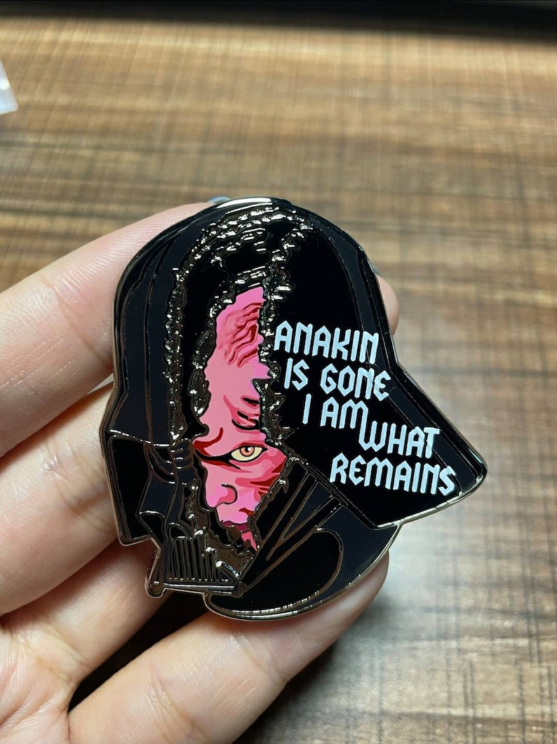 May include: A black enamel pin featuring Darth Vader's helmet with a split revealing Anakin's face. The text "ANAKIN IS GONE I AM WHAT REMAINS" is displayed in white. The pin is a collectible item.