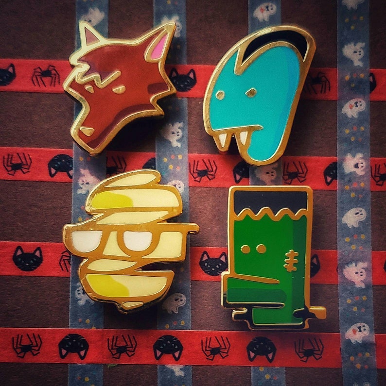 May include: Four enamel pins featuring Halloween characters. A red wolf, a blue monster with fangs, a yellow mummy with glasses, and a green Frankenstein's monster.