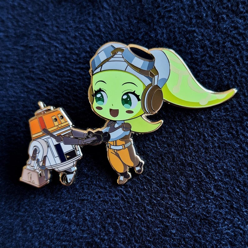 May include: Enamel pin featuring a green-skinned character with a yellow ponytail, wearing goggles and a brown and orange outfit, holding a blaster and standing next to a small orange and white droid. The pin is set against a dark blue background.