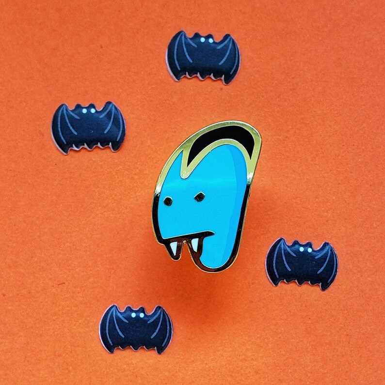 May include: A blue and gold enamel pin of a vampire with fangs. The pin is surrounded by five black enamel pins shaped like bats.