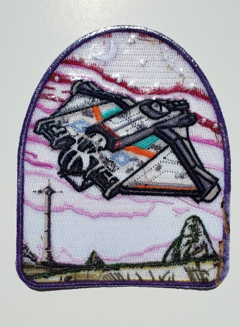 May include: Embroidered patch featuring a white spaceship with orange and teal accents flying over a pink and purple landscape. The patch has a purple border and is sewn onto a white background.