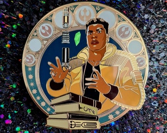 Può includere: Una spilla in smalto dorata con un personaggio di Star Wars. Il personaggio è un giovane uomo con la pelle scura, che indossa una giacca gialla e una felpa con cappuccio nera. Tiene una spada laser nella mano destra e un mucchio di libri nella mano sinistra. La spilla è circondata da un design circolare con sfondo blu e accenti dorati.