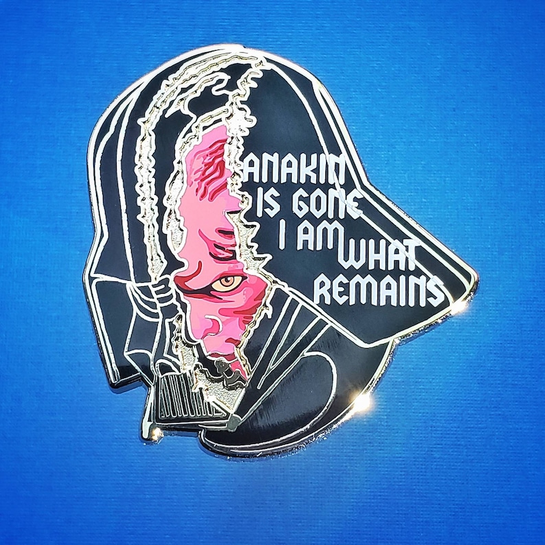 May include: A black and silver enamel pin featuring a split design of Darth Vader and Anakin Skywalker. The text "ANAKIN IS GONE I AM WHAT REMAINS" is displayed in white. The pin has a shiny, metallic finish.