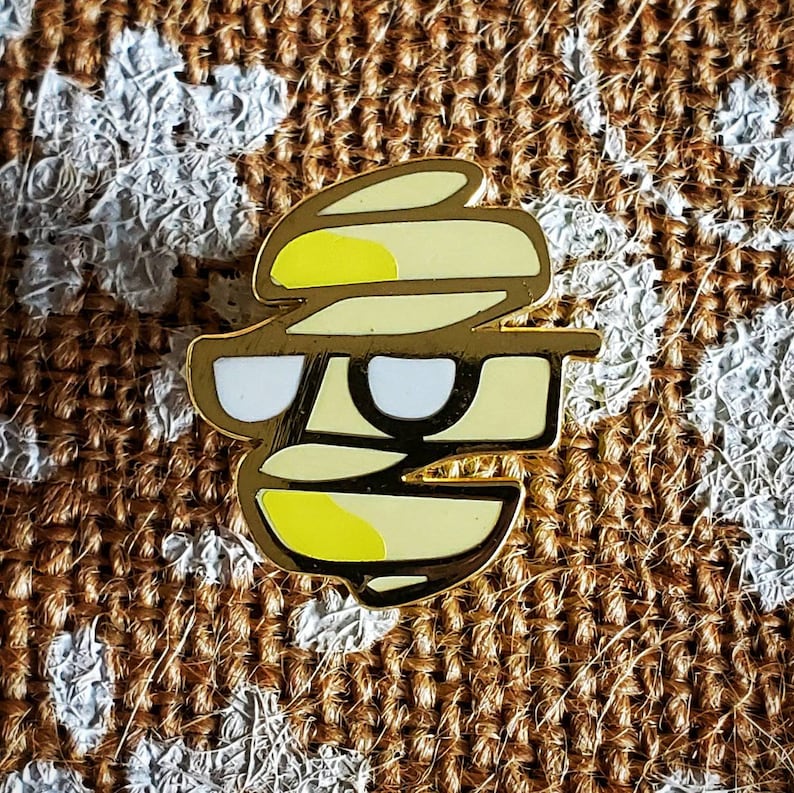 May include: A gold-colored enamel pin with a yellow and white design. The design is a stylized face with sunglasses and a layered hat.
