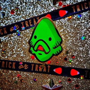 May include: Green enamel pin with a cartoon monster design. The monster has a large mouth with red lips and a green body with a white outline. The pin is on a sparkly gold background with a black and red ribbon that says "Trick or Treat".
