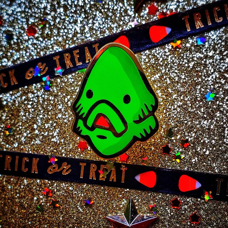 May include: Green enamel pin with a cartoon monster design. The monster has a large mouth with red lips and a green body with a white outline. The pin is on a sparkly gold background with a black and red ribbon that says "Trick or Treat".