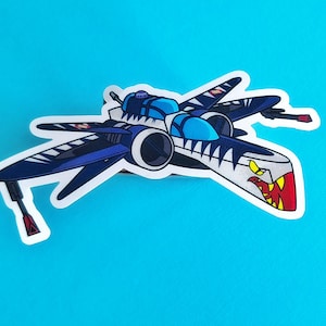 May include: A sticker of a cartoon spaceship in shades of blue, white, and black. The spaceship has a stylized shark mouth design on the front. The background is a solid light blue.
