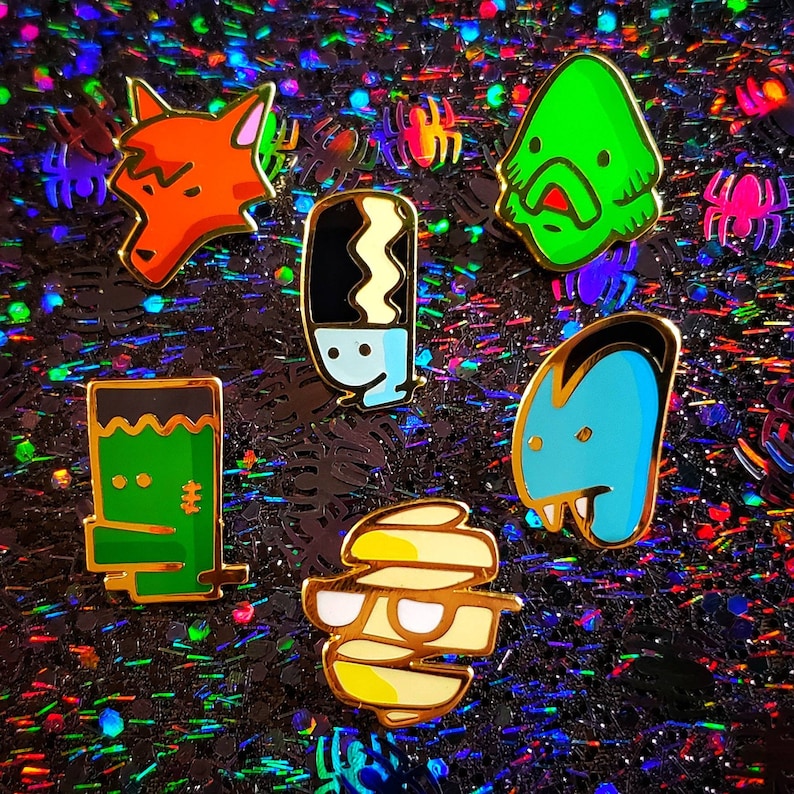 May include: A set of six enamel pins featuring various Halloween characters. The pins are gold-toned and feature a mummy, a monster, a werewolf, a ghost, a creature from the black lagoon, and a fox.