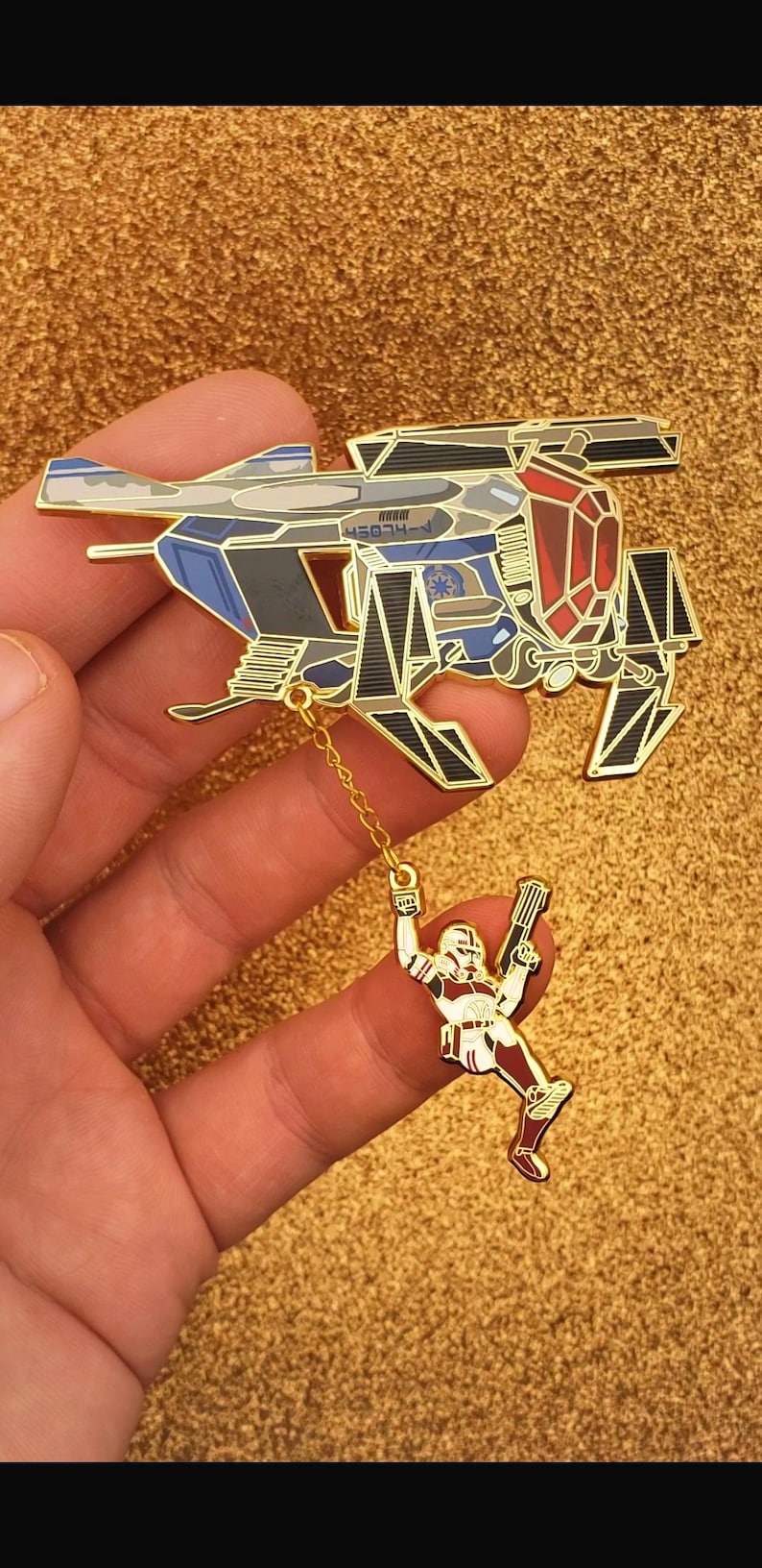 May include: A gold-colored pin featuring a detailed spaceship design with blue, gray, and red accents. A small figure of a soldier hangs from the ship by a chain. The pin is set against a gold background.