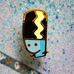 May include: Gold-tone enamel pin featuring a cartoon character with a black and yellow striped hair, blue eyes, and a light blue body. The character is smiling.