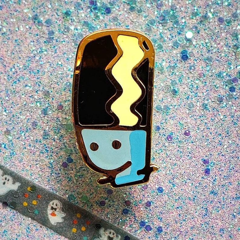 May include: Gold-tone enamel pin featuring a cartoon character with a black and yellow striped hair, blue eyes, and a light blue body. The character is smiling.