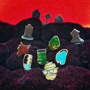 May include: A set of eight enamel pins featuring various Halloween characters, including a werewolf, a mummy, a ghost, a vampire, and a Frankenstein's monster. The pins are arranged on a black fuzzy surface with miniature tombstones in the background.