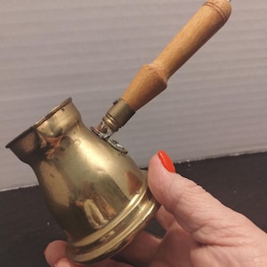 May include: A vintage brass coffee pot with a wooden handle and a hook. The pot has a rounded base and a flared top. The handle is attached with a metal band. The pot is held in a hand with red nail polish.