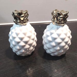 May include: Two white ceramic pineapple-shaped decorative objects with gold-colored tops. The pineapples have a textured, geometric pattern. The objects are displayed on a dark surface.