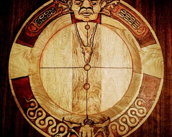 May include: A circular wooden platter with a carved design. The center of the platter features a cross with a human face at the top and a deer head at the bottom. The outer edge of the platter is decorated with snakes and other intricate carvings.