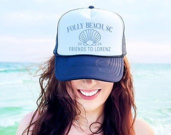 May include: A woman wearing a black and white trucker hat with the text "FOLLY BEACH, SC 20124 FRIENDS TO LORENZ" and a seashell graphic. The hat is worn backwards and the woman is smiling.