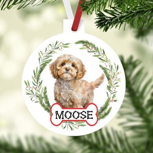 May include: A white round Christmas ornament with a watercolor illustration of a brown and white dog. The dog is surrounded by a green wreath with red berries. The ornament has a red bone-shaped tag with the name "MOOSE" written in black.