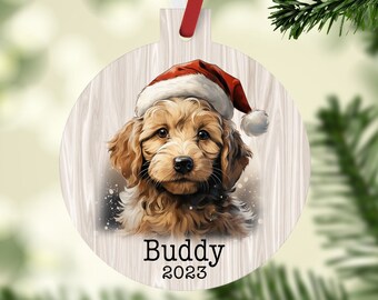 May include: Round Christmas ornament featuring a watercolor illustration of a golden doodle puppy wearing a Santa hat. The ornament has the name "Buddy" and the year "2023" printed on it. The ornament hangs from a red and white ribbon.