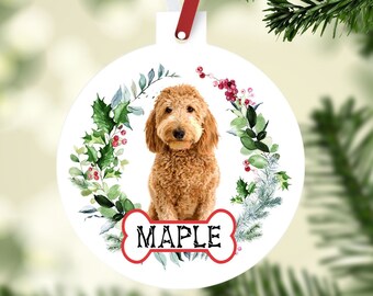 May include: A white round Christmas ornament with a watercolor illustration of a brown and white dog sitting in a wreath of green leaves and red berries. The dog's name, "Maple", is written on a white bone-shaped tag.