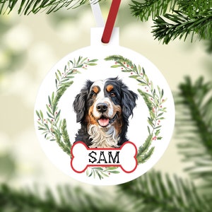 May include: White round Christmas ornament with a watercolor illustration of a Bernese Mountain Dog with a red and white bone with the name "SAM" on it. The dog is surrounded by a green wreath with red berries.
