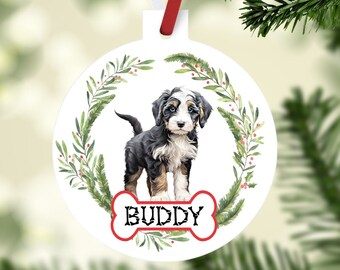 May include: White circular Christmas ornament with a Bernedoodle puppy illustration. The puppy is black, white, and brown, with the name "BUDDY" on a bone-shaped design. The ornament is decorated with a green wreath and red berries. A red and white ribbon is attached.