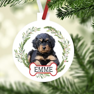 May include: White round Christmas ornament featuring a puppy with black and tan fur, surrounded by a green wreath. The name "EMME" is displayed on a bone-shaped graphic. The ornament hangs from a red and white ribbon.