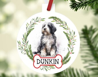 May include: White round Christmas ornament featuring a watercolor illustration of a dog with black and gray fur, surrounded by a green wreath with red berries. The dog's name, "Dunkin," is displayed on a bone-shaped tag.