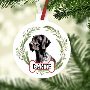 Peut inclure: Ornement de Noël rond blanc avec un portrait de chien Pointer allemand, encadré d'une couronne verte avec des baies rouges. Le nom du chien, "DANTE", est imprimé sur un design en forme d'os.