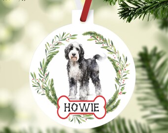 May include: White round Christmas bauble featuring a watercolour illustration of a black and white dog with a red bone and the name "HOWIE" on it. The bauble is surrounded by a green wreath with red berries.