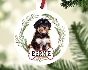 May include: White round Christmas ornament featuring a puppy with black and brown fur, a red bone-shaped name tag that says "Bernie", and a green wreath. The ornament hangs from a red and white ribbon.