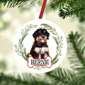 May include: White round Christmas ornament featuring a puppy with black and brown fur, a red bone-shaped name tag that says "Bernie", and a green wreath. The ornament hangs from a red and white ribbon.