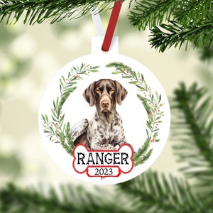 May include: White round Christmas ornament with a brown and white German Shorthaired Pointer dog illustration. The dog is sitting with a green wreath around it. The ornament has a bone-shaped tag with the text "RANGER 2023".