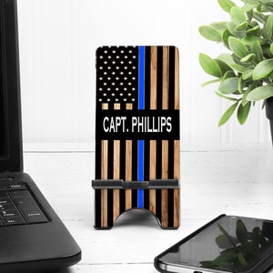 May include: A black phone case with a thin blue line and an American flag design. The case has a kickstand and says "CAPT. PHILLIPS" in white text.